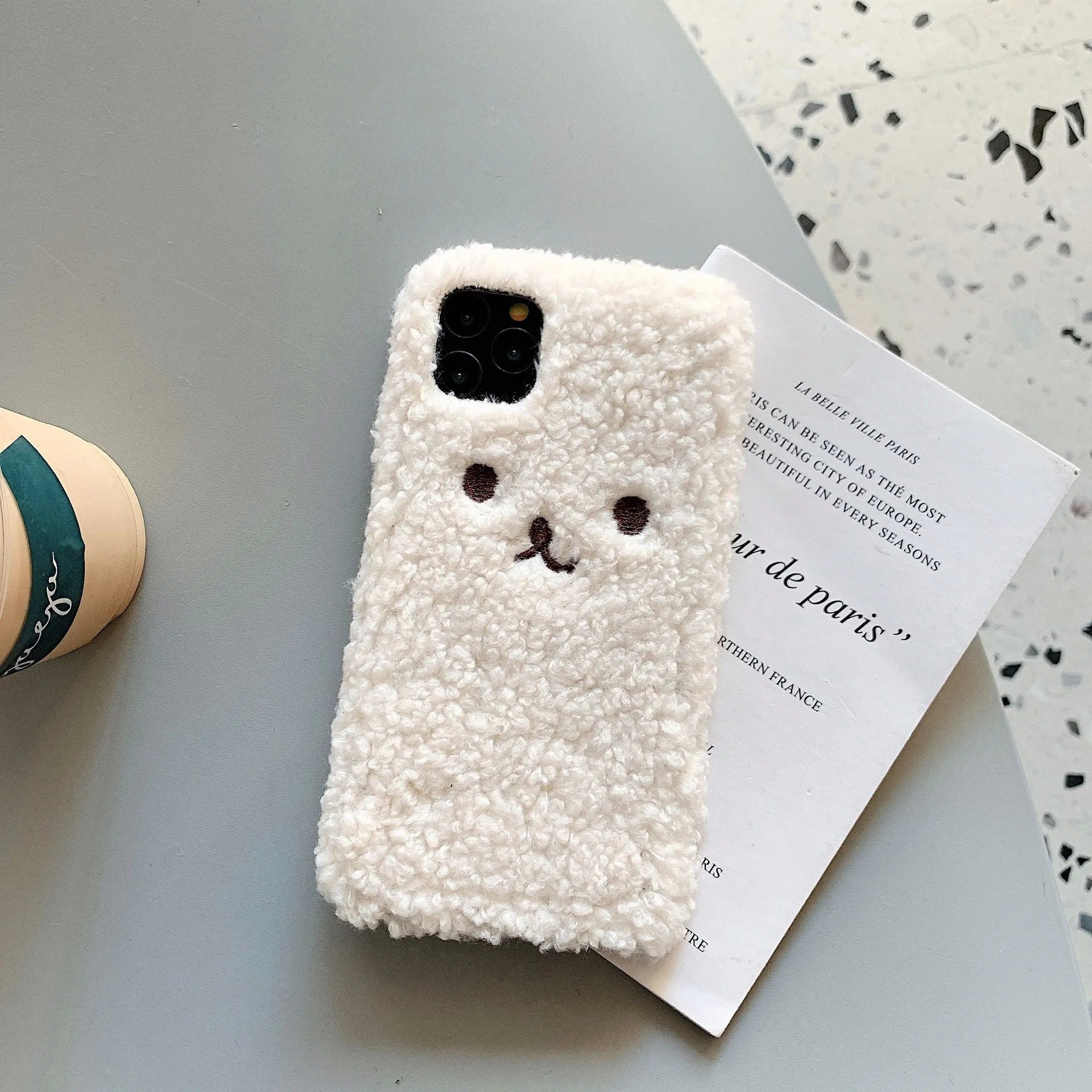 Brown embroidered plush bear face on a soft TPU phone case for iPhone, showing camera cut-out and raised edges.