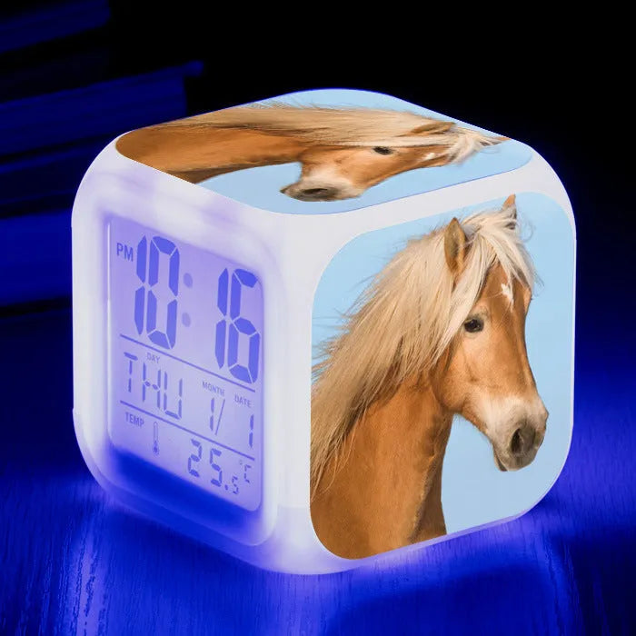Small square colour changing alarm clock with LED digital display on a desk.