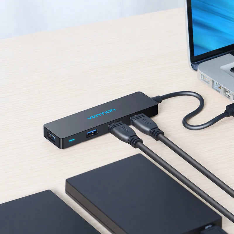 Black four-port USB 3.0 hub with a short, thick cable on a wooden desk next to a laptop.
