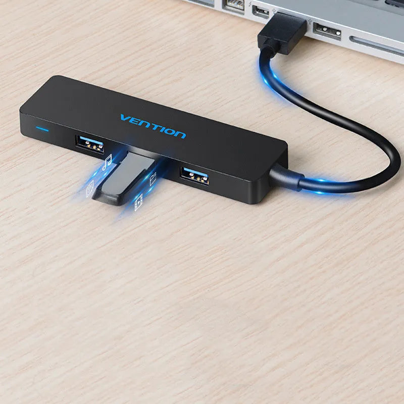 Black four-port USB 3.0 hub with a short, thick cable on a wooden desk next to a laptop.