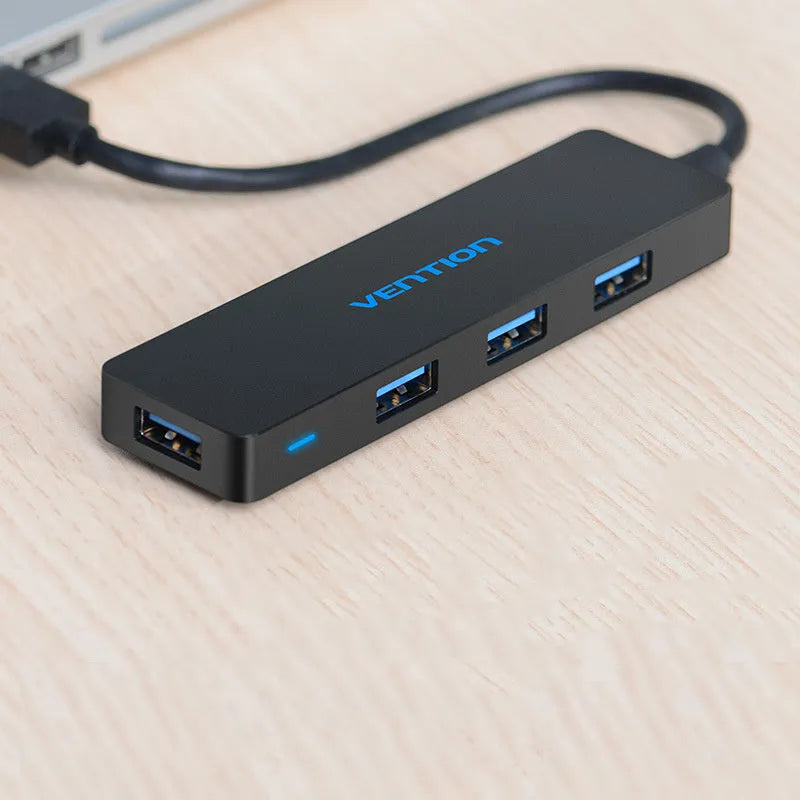 Black four-port USB 3.0 hub with a short, thick cable on a wooden desk next to a laptop.