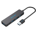 Black four-port USB 3.0 hub with a short, thick cable on a wooden desk next to a laptop.