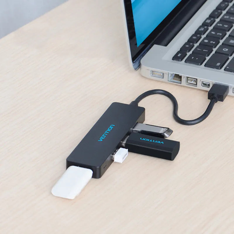 Black four-port USB 3.0 hub with a short, thick cable on a wooden desk next to a laptop.