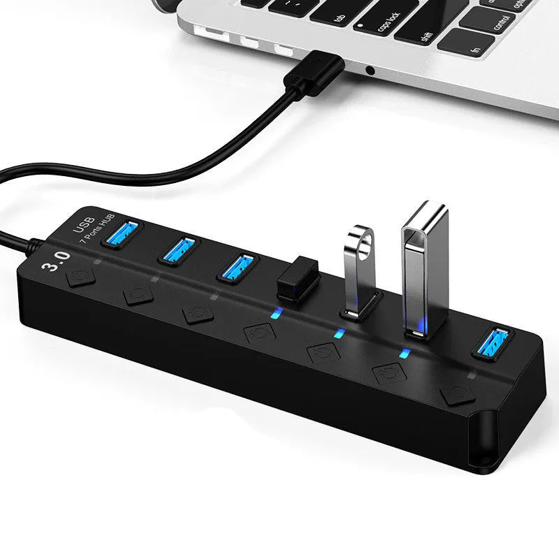 Black 7-port USB 3.0 hub with individual power switches and external power adapter on a wooden desk.
