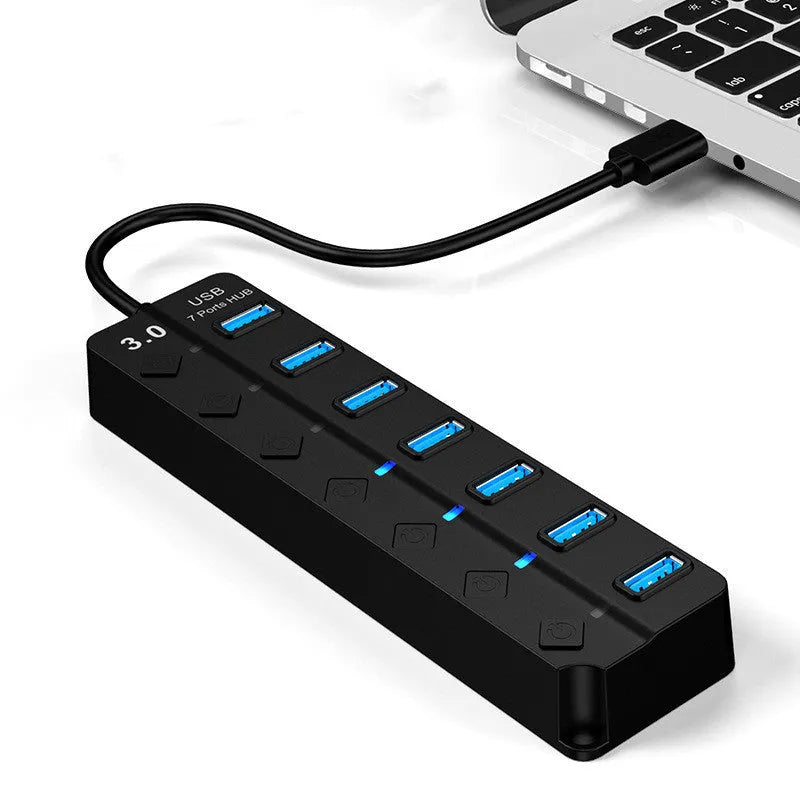 Black 7-port USB 3.0 hub with individual power switches and external power adapter on a wooden desk.