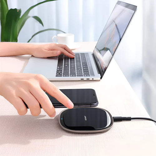 Black 15W wireless charging pad with glass top on a wooden desk next to a mobile phone.
