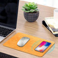 Brown leather wireless charging mouse pad on a wooden desk with a smartphone charging on its surface.