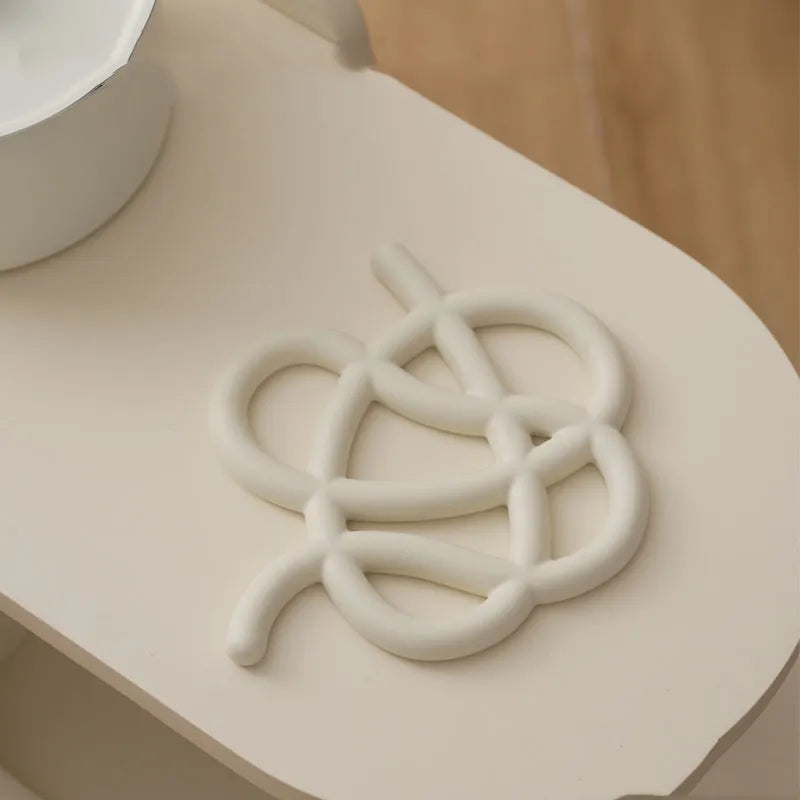 Irregular-shaped ceramic heat proof mat in grey with a curved hook for hanging, sitting on a wooden table.