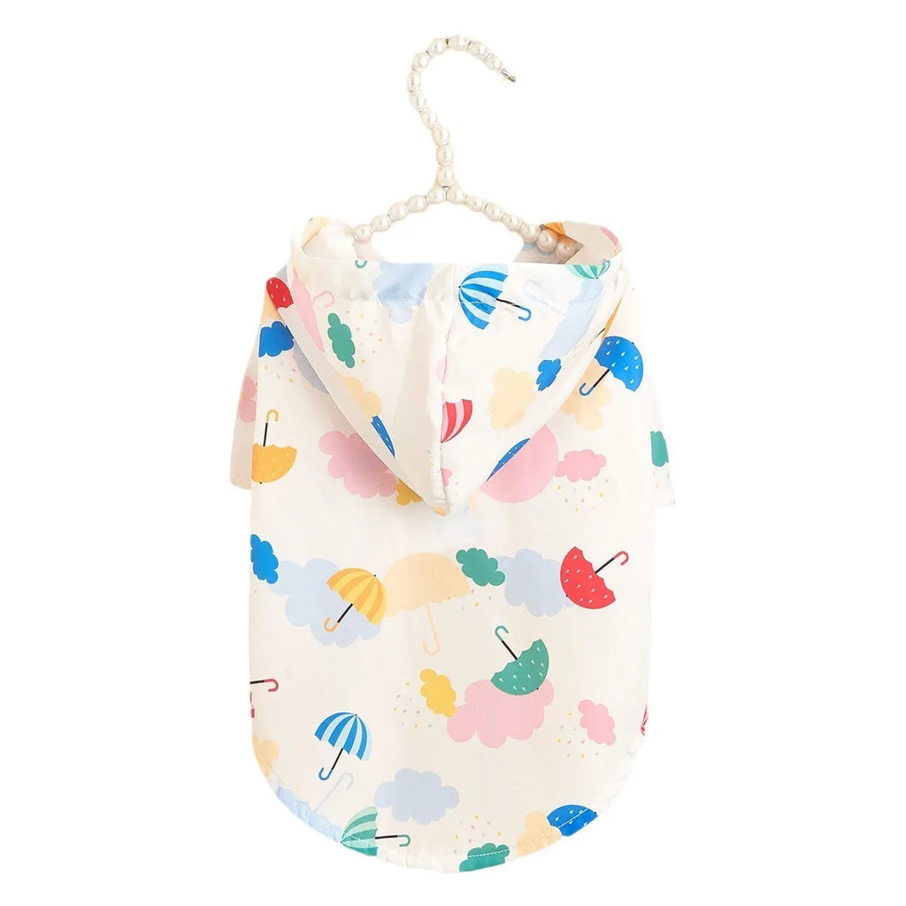 Small white dog wearing a colourful cartoon umbrella pattern polyester raincoat outdoors.