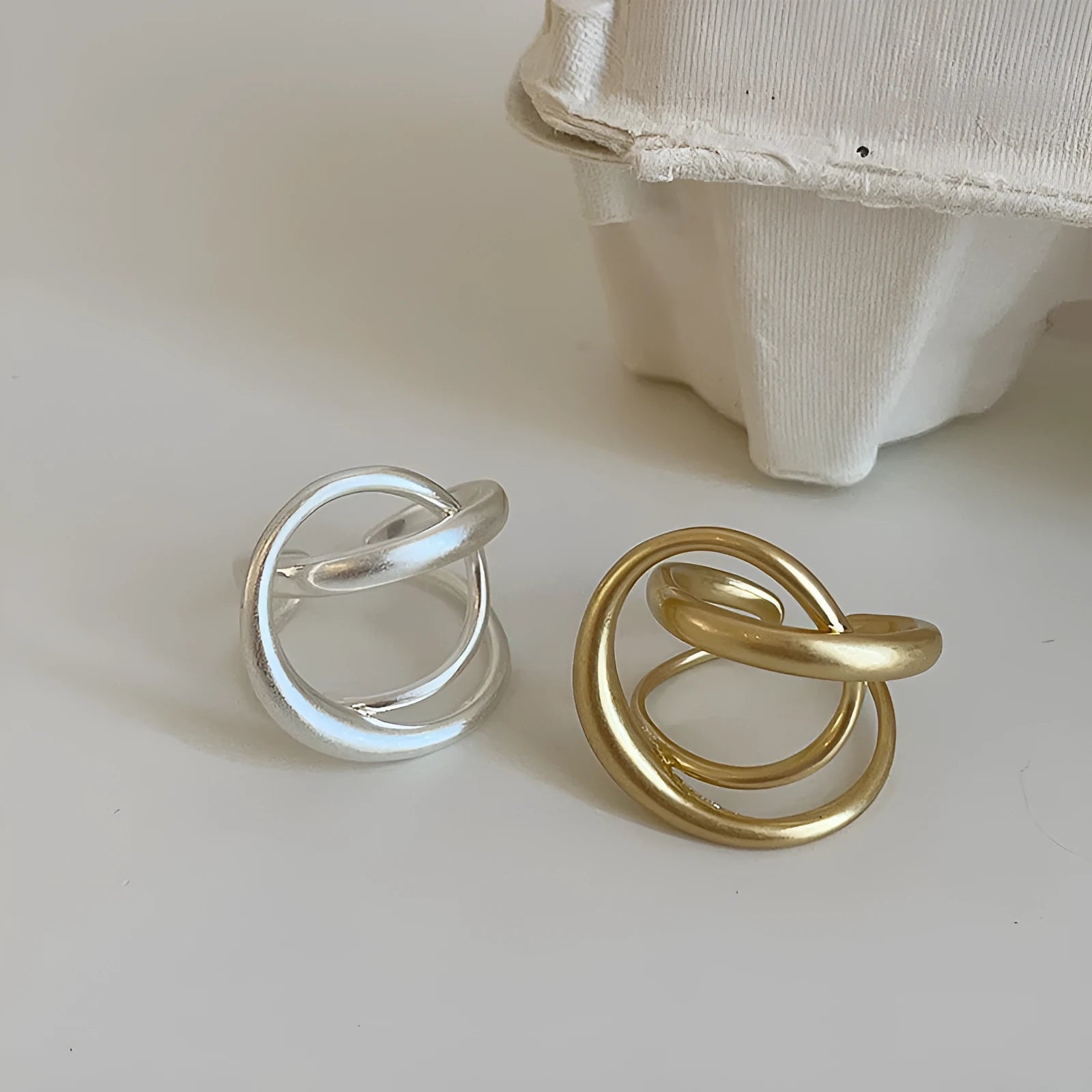 Geometric open ring with a 3D interlaced design in matte gold or silver metal.