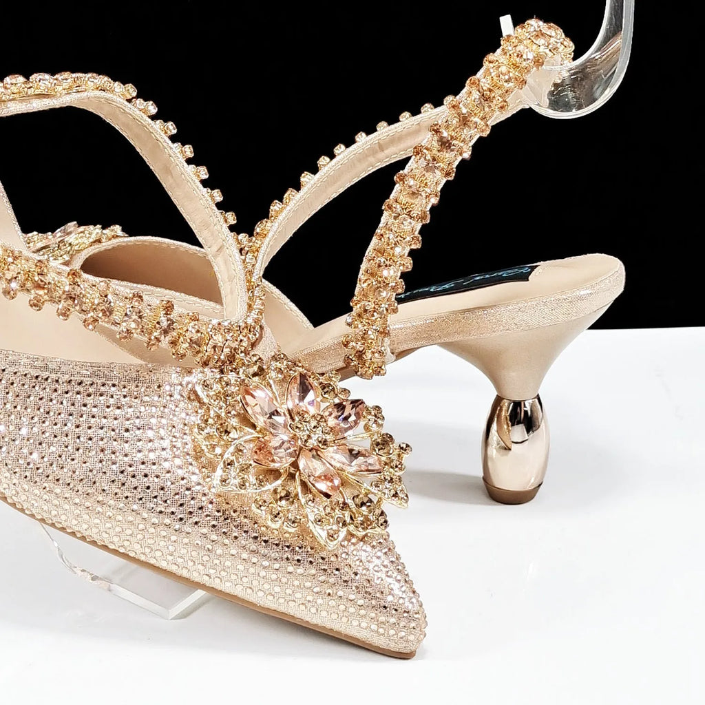 Women's black high heel shoes with rhinestone flowers and an ankle chain, next to a matching hard clutch bag.
