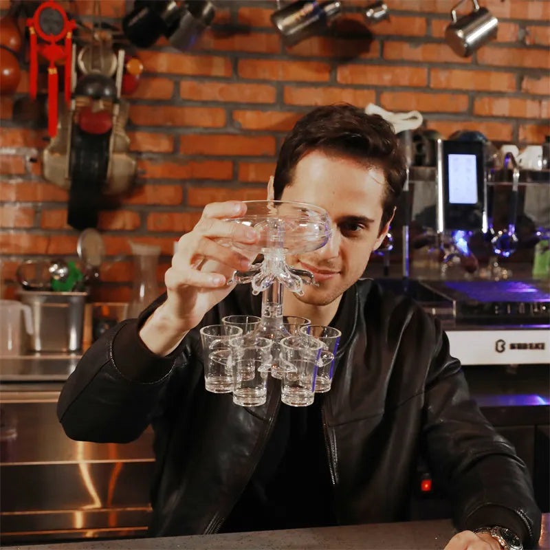 Clear six-shot glass dispenser holder with rack and six cups on a bar table.