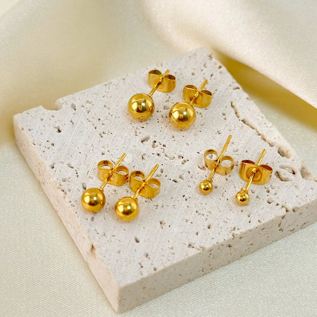Close-up of a simple gold ball stud earring on a titanium steel post against a plain background.