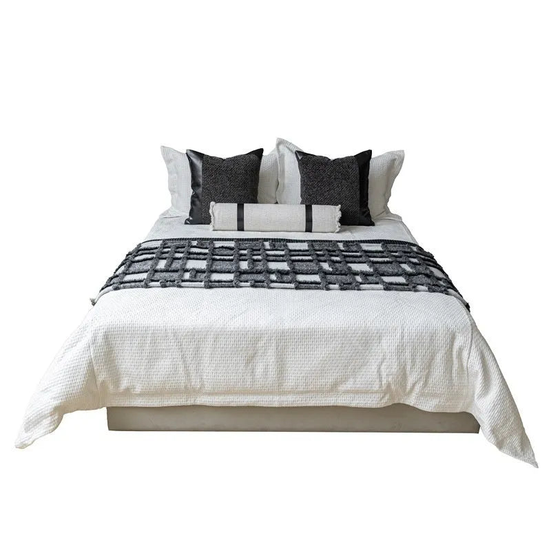 Grey and white plaid bed runner blanket with matching pillowcase and brown leather round pillow on a bed.