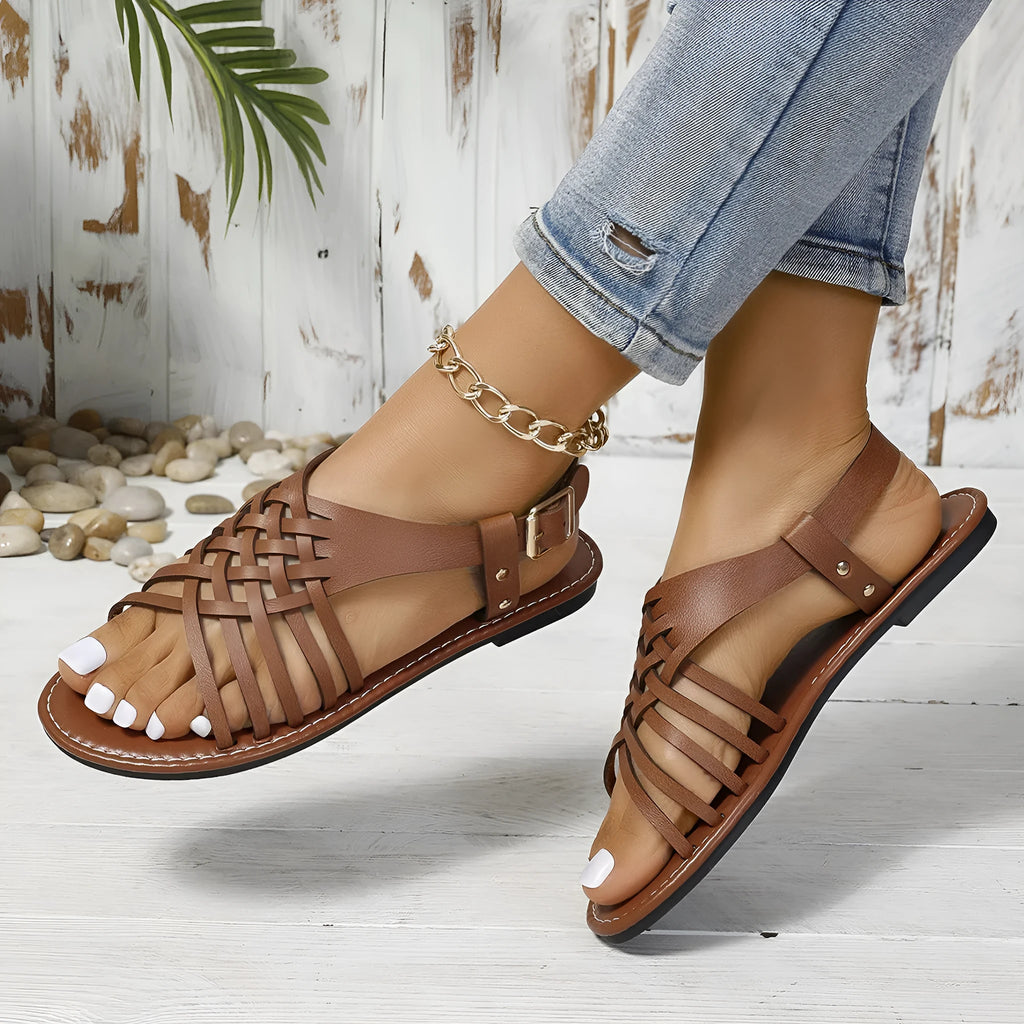 Flat buckle open toe Roman style cross strap sandals in black on a beige background.