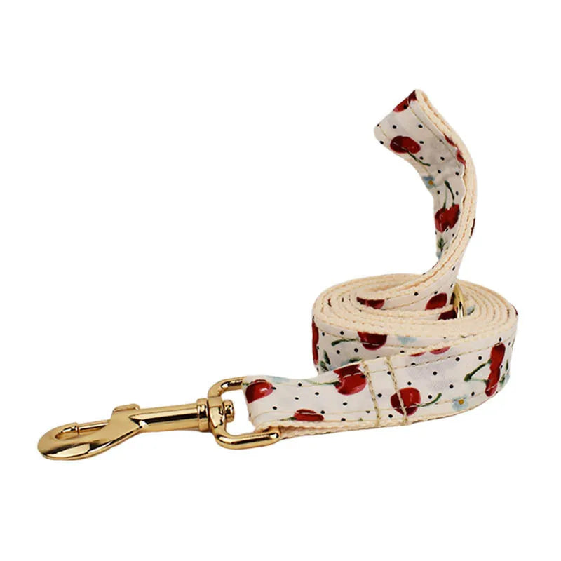 Red cherry print pet collar, lead, and harness set with light gold buckles and matching waste bag dispenser