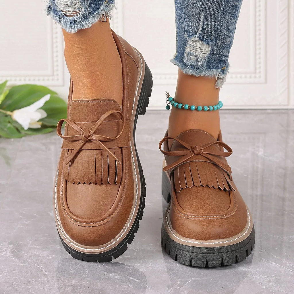Women's black loafers with a tassel and bow detail on a chunky rubber sole.