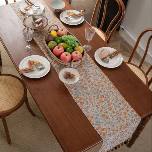 Long beige jacquard table runner with floral pattern and tassels draped on a wooden dining table.