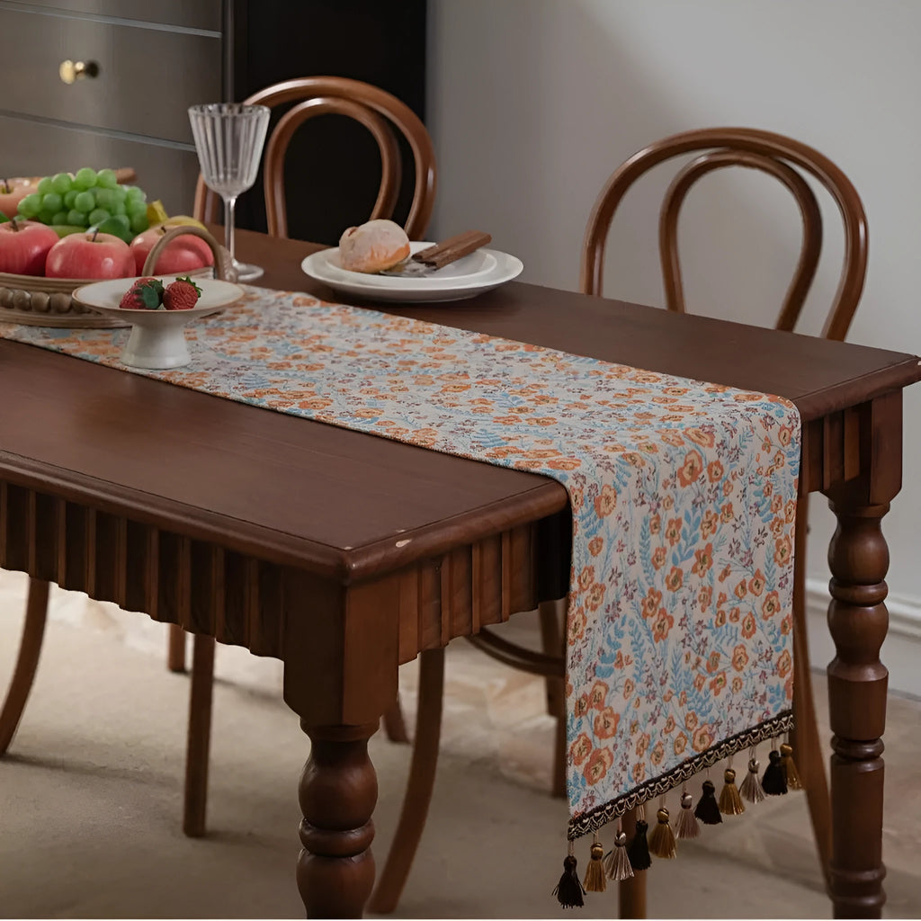 Long beige jacquard table runner with floral pattern and tassels draped on a wooden dining table.