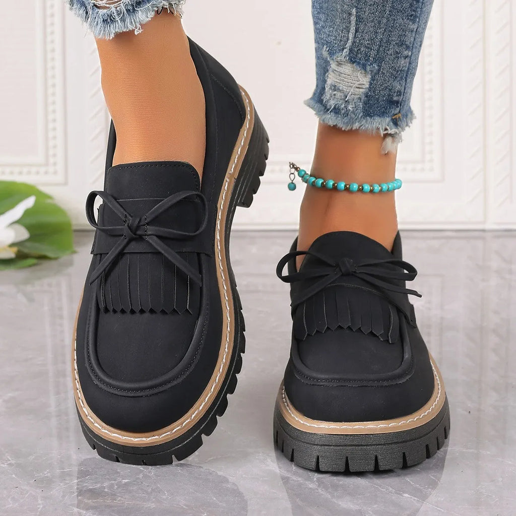 Women's black loafers with a tassel and bow detail on a chunky rubber sole.