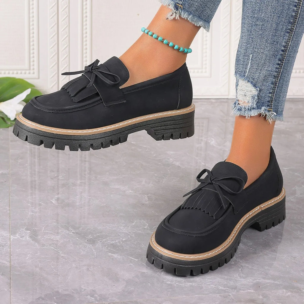 Women's black loafers with a tassel and bow detail on a chunky rubber sole.