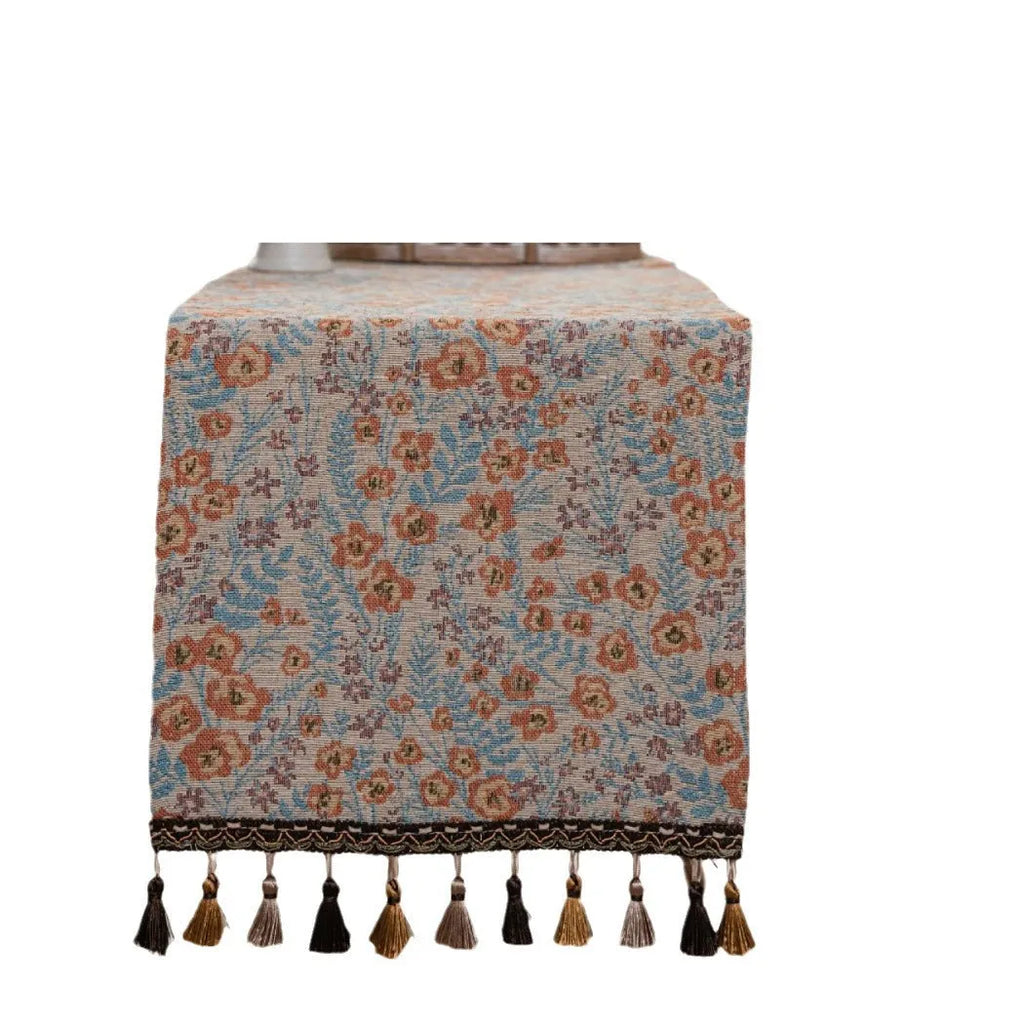 Long beige jacquard table runner with floral pattern and tassels draped on a wooden dining table.