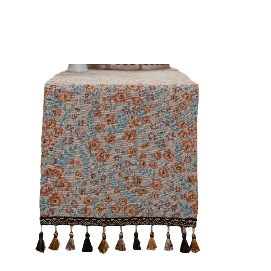 Long beige jacquard table runner with floral pattern and tassels draped on a wooden dining table.