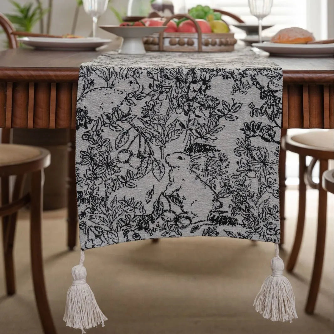 Long beige jacquard table runner with floral pattern and tassels draped on a wooden dining table.