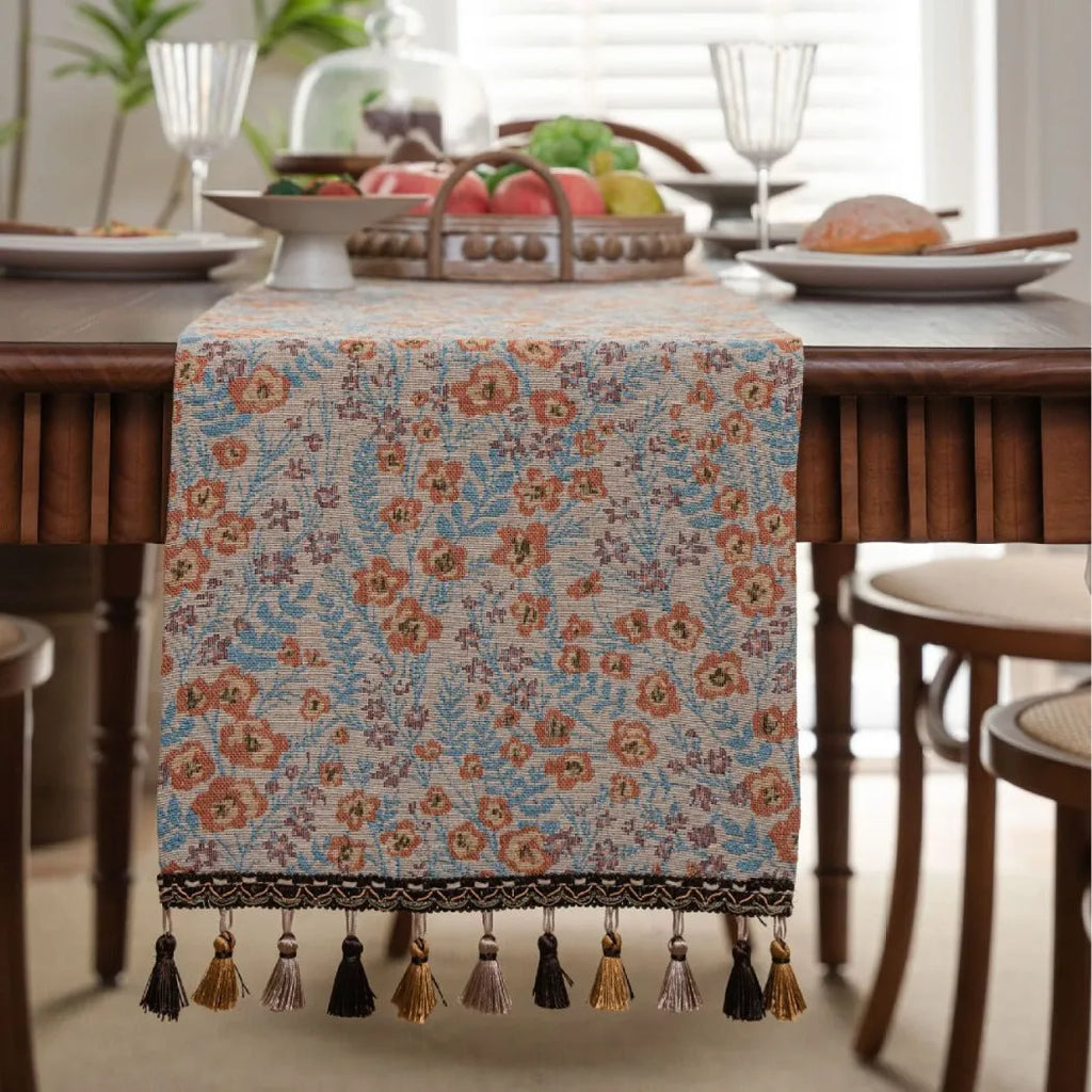 Long beige jacquard table runner with floral pattern and tassels draped on a wooden dining table.