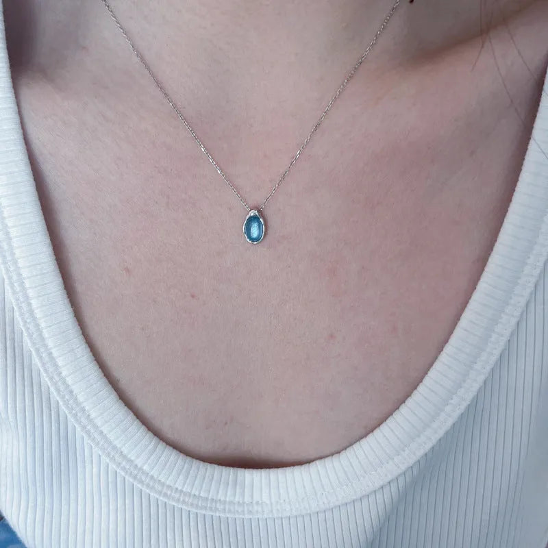 Close-up of a navy blue and platinum coloured water drop pendant necklace with an adjustable cross chain on a person's collarbone.