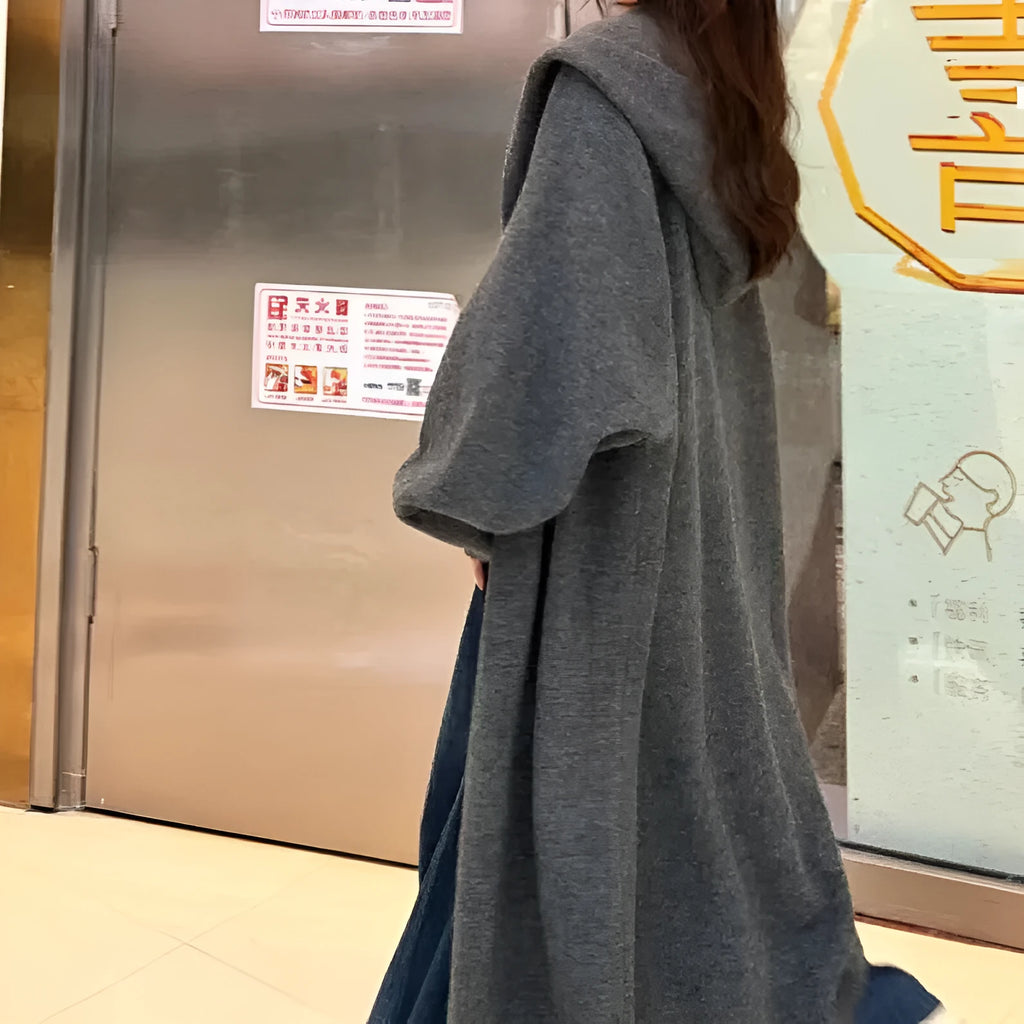 A woman wearing a long, beige, hooded knitted cardigan in a loose, below-the-knee Korean style.