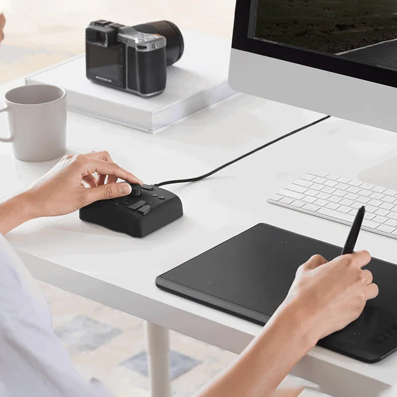 TourBox NEO black video editing controller with dials and buttons on a desk next to a tablet.