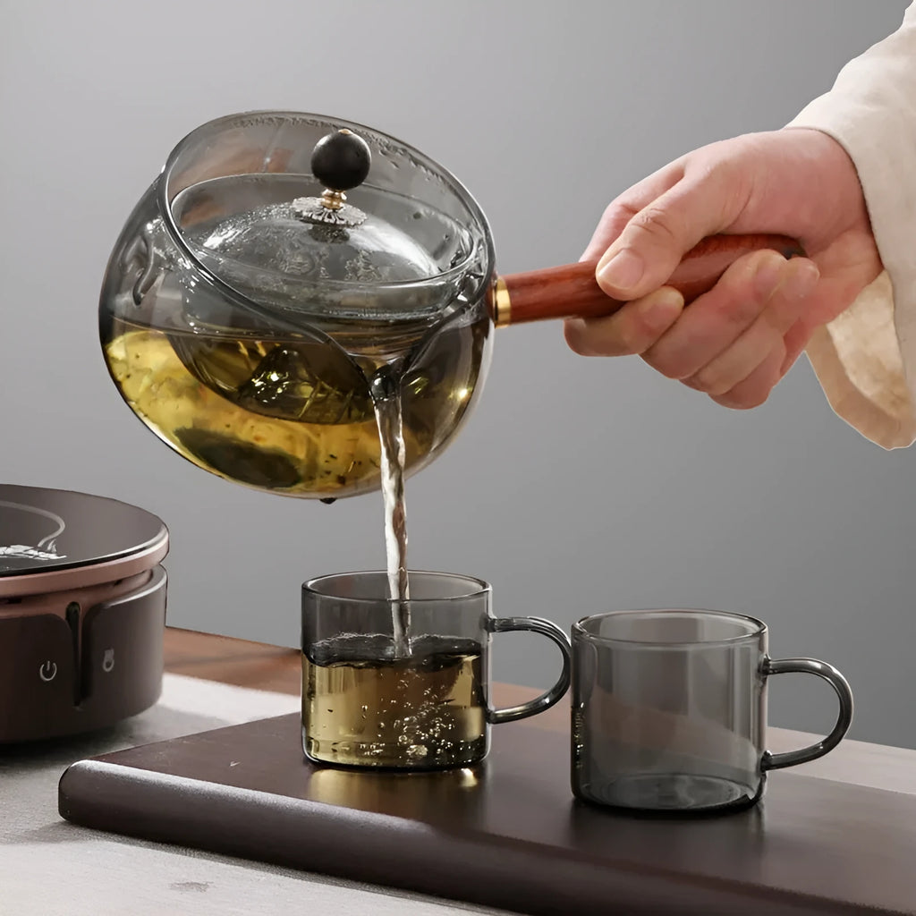 Grey and transparent rotary glass teapot with wooden handle and infuser on a kitchen counter