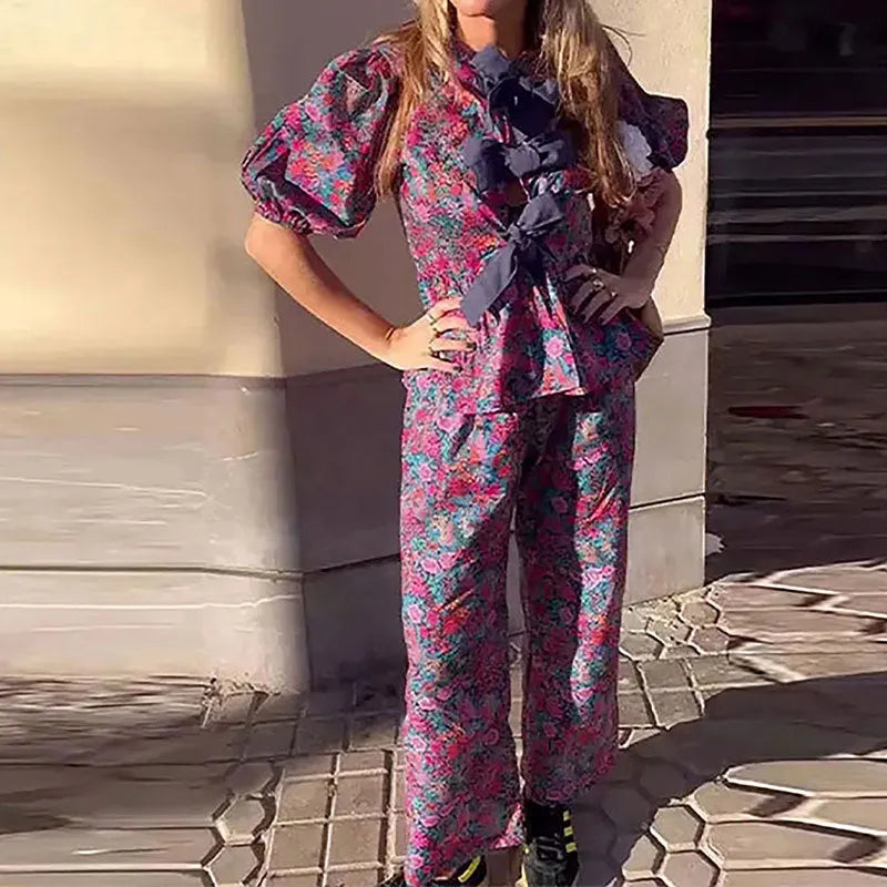 Women's floral print polyester trousers suit with tie-neck blouse and puff sleeves.