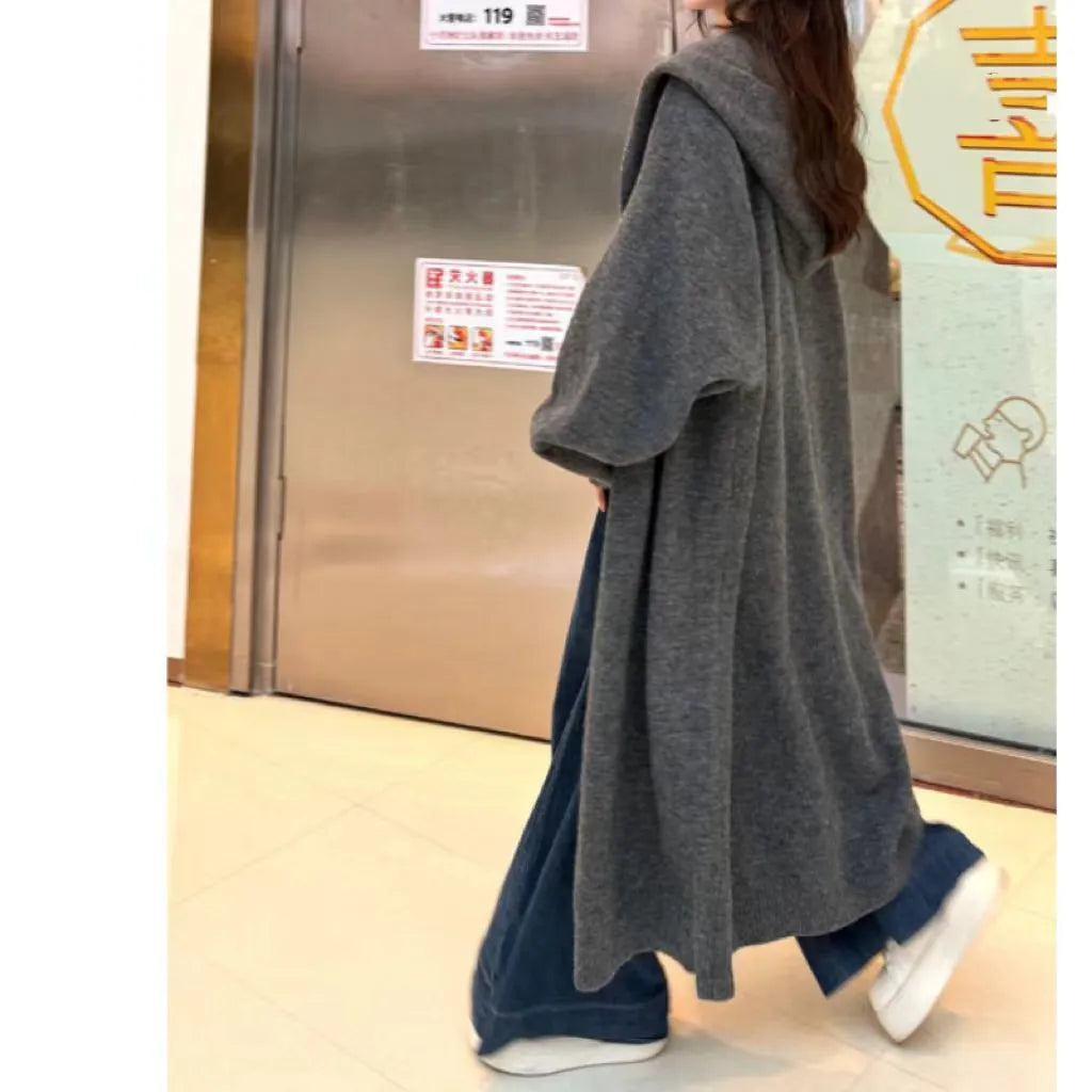 A woman wearing a long, beige, hooded knitted cardigan in a loose, below-the-knee Korean style.