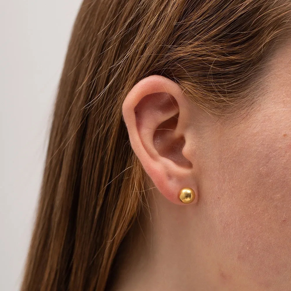 Close-up of a simple gold ball stud earring on a titanium steel post against a plain background.