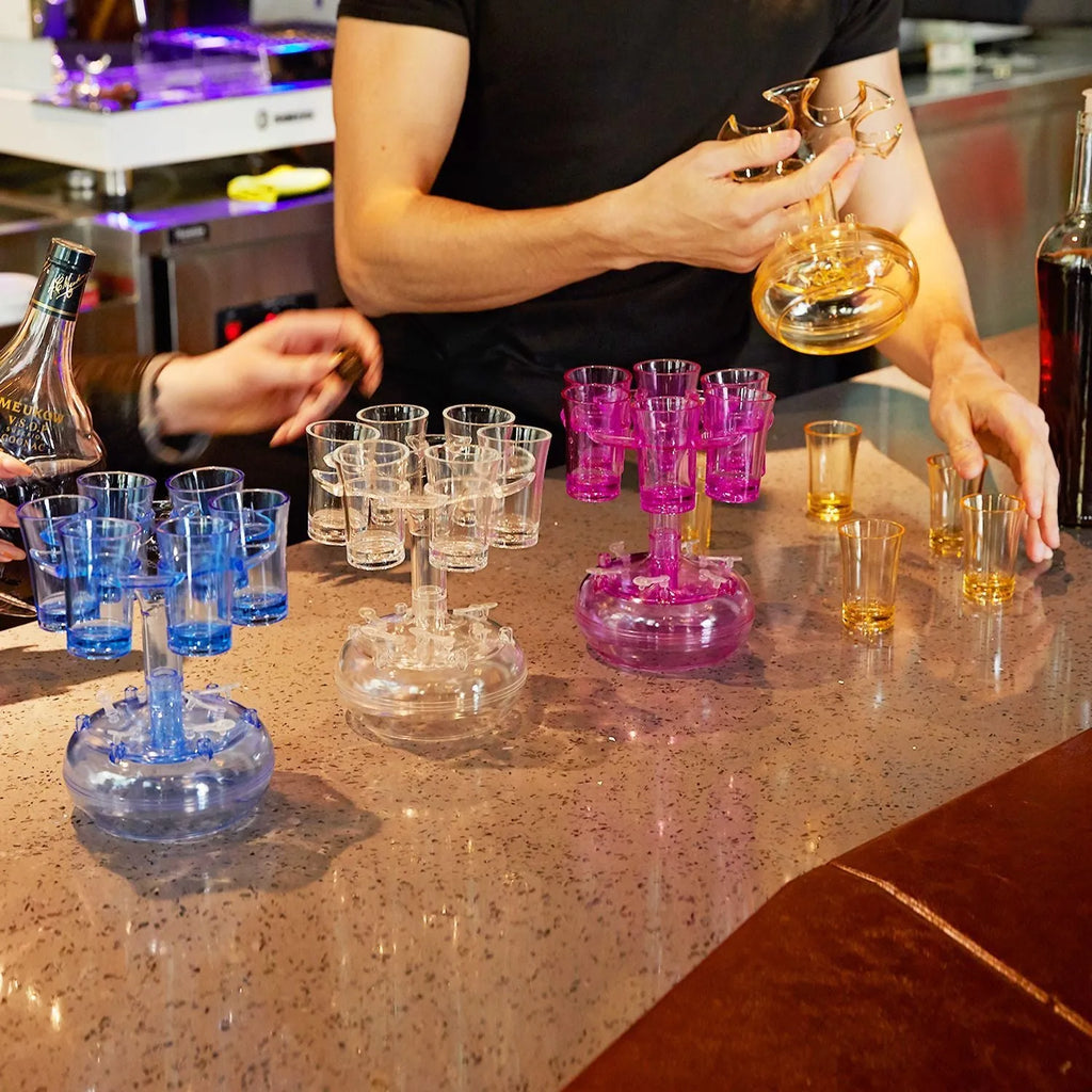 Clear six-shot glass dispenser holder with rack and six cups on a bar table.